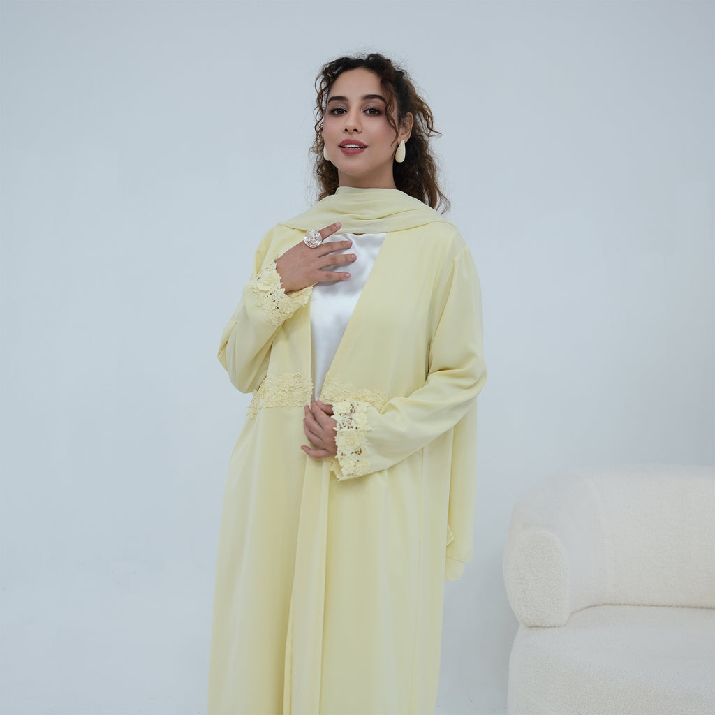 A woman models a pale yellow long abaya with matching scarf and a white inner top, standing against a light gray studio background.
