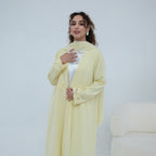A woman models a pale yellow long abaya with matching scarf and a white inner top, standing against a light gray studio background.