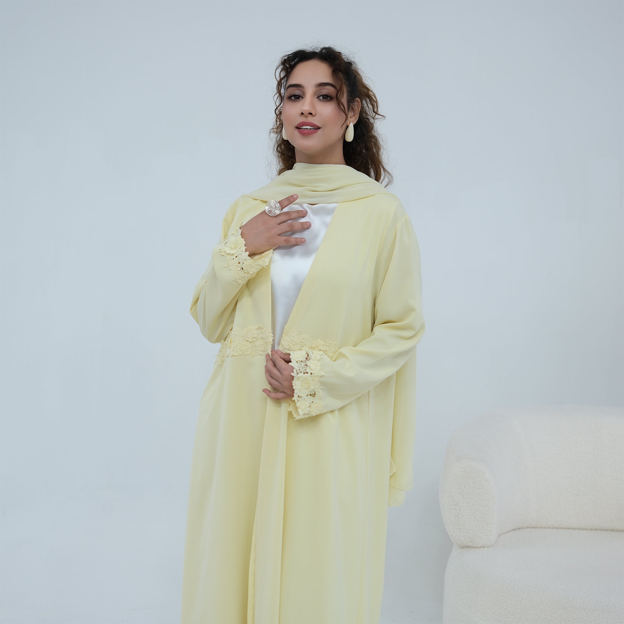 A woman models a pale yellow long abaya with matching scarf and a white inner top, standing against a light gray studio background.