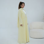A woman models a long, flowing pale yellow abaya dress with lace trim details, standing in a bright, minimalist studio setting with a cream-colored armchair in the background.