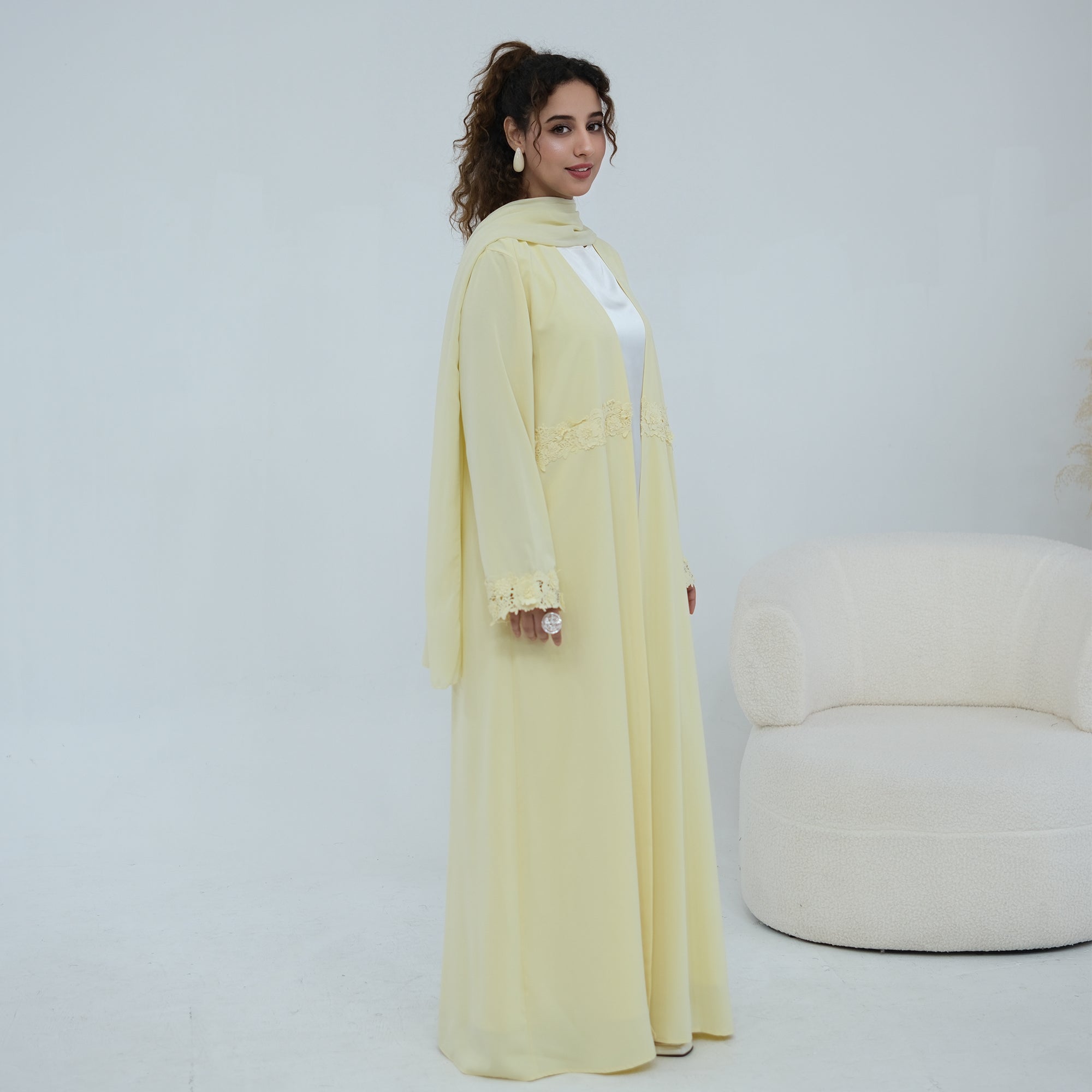 A woman models a long, flowing pale yellow abaya dress with lace trim details, standing in a bright, minimalist studio setting with a cream-colored armchair in the background.