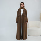 A woman models a long, flowing brown abaya with matching headscarf, worn over a white inner dress. The abaya features delicate lace trim details at the waist and cuffs.