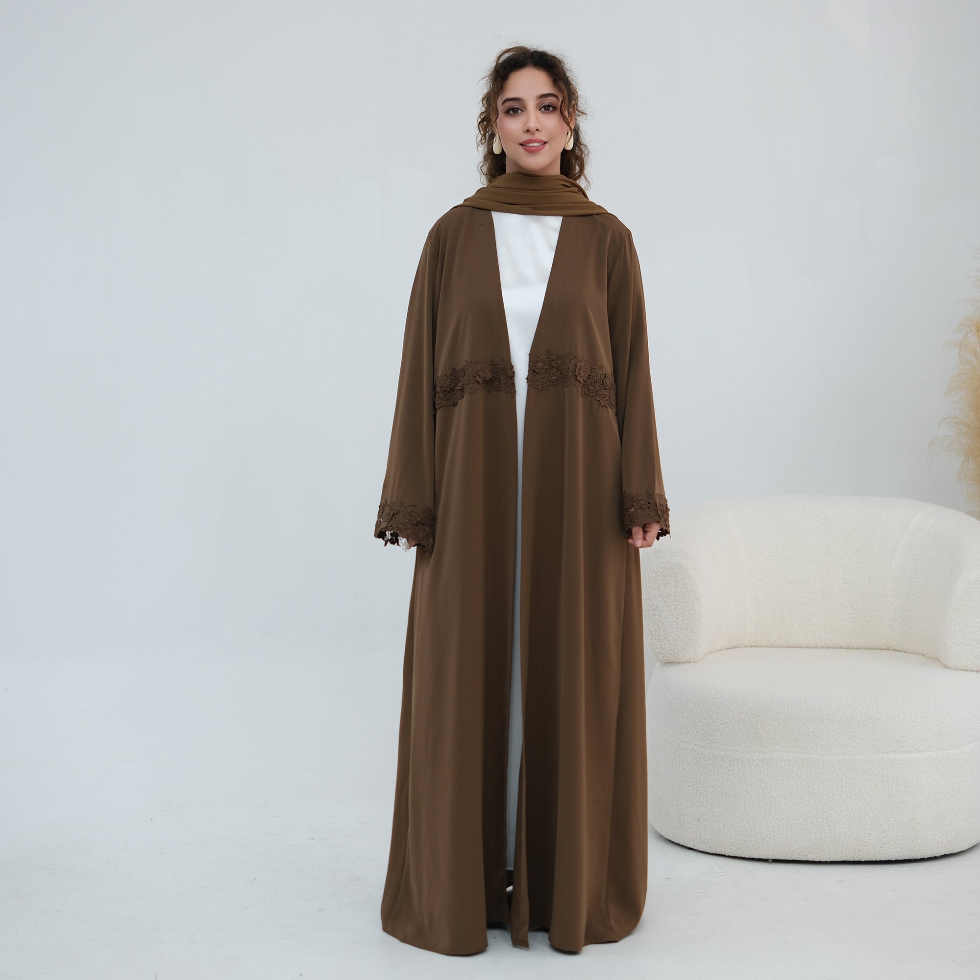 A woman models a long, flowing brown abaya with matching headscarf, worn over a white inner dress. The abaya features delicate lace trim details at the waist and cuffs.