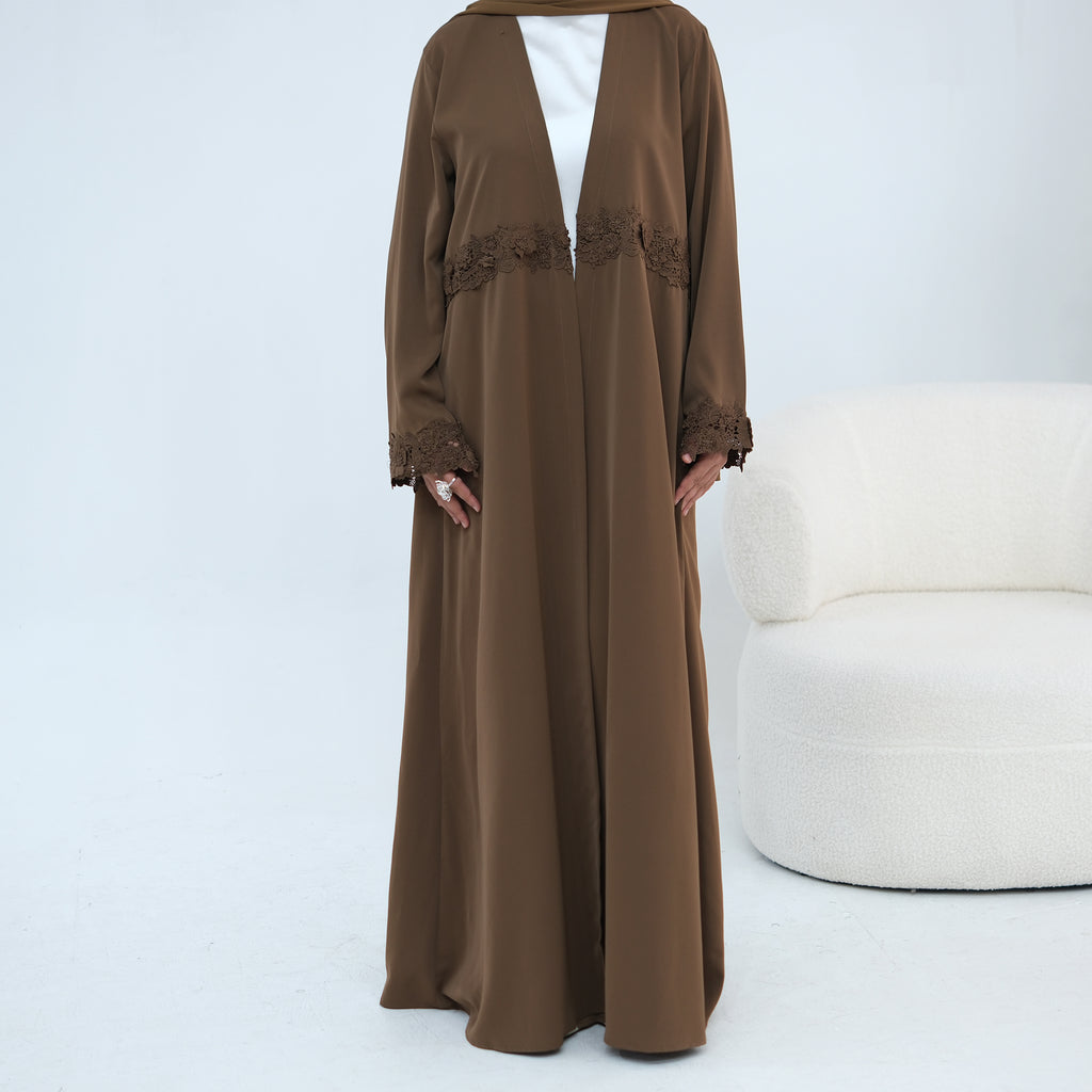 A woman modeling a long brown abaya dress with lace trim details, worn over a white top, standing in a studio setting with a white textured chair on the right.