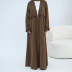 A woman modeling a long brown abaya dress with lace trim details, worn over a white top, standing in a studio setting with a white textured chair on the right.