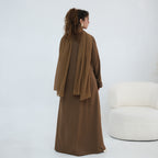 A woman stands with her back to the camera wearing a long brown abaya with a draped shawl, posed next to a modern white armchair in a minimalist studio setting.