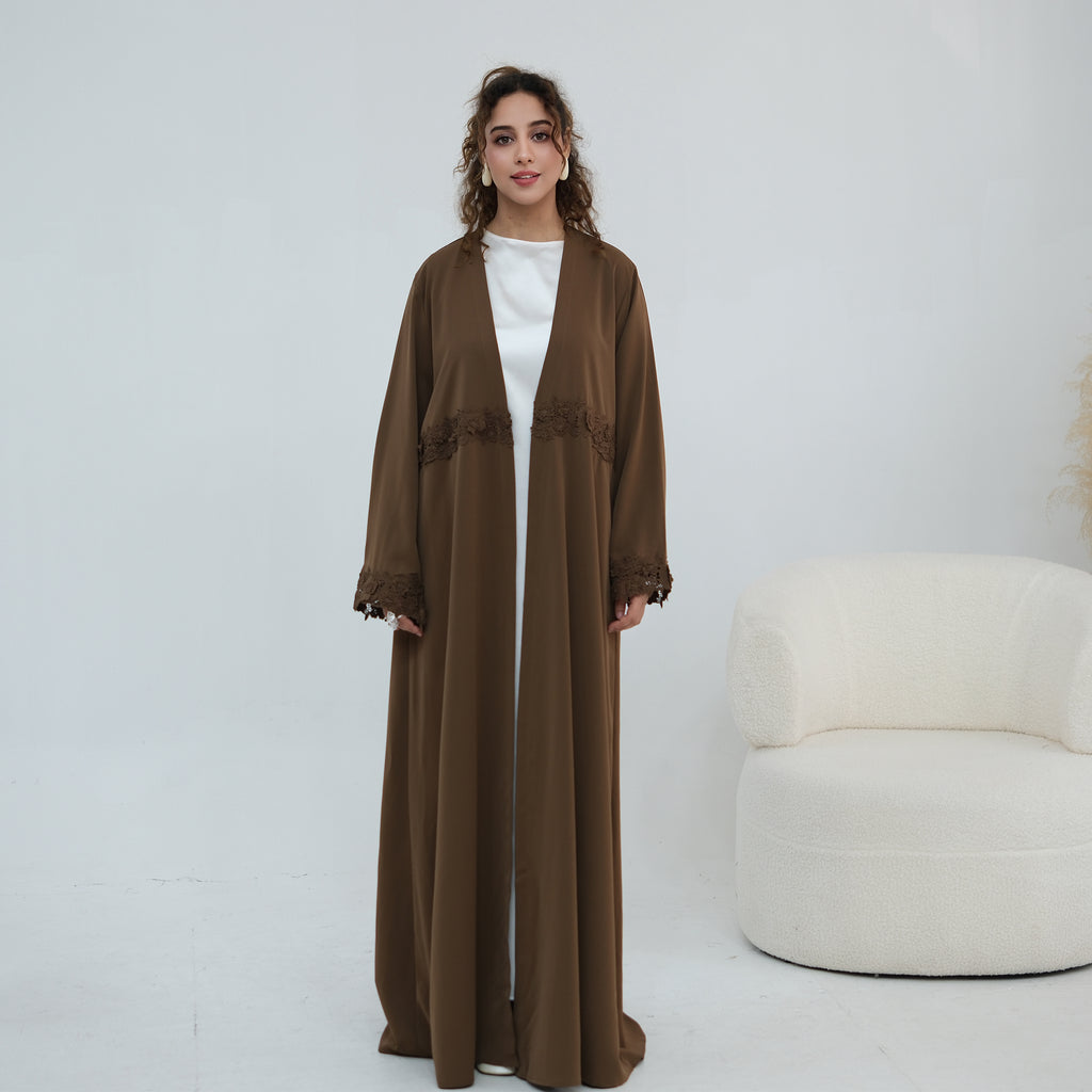 A woman stands in a studio wearing a long brown open-front abaya or kaftan with lace trim details, layered over a white top. A white curved armchair is visible to the right against a plain light background.