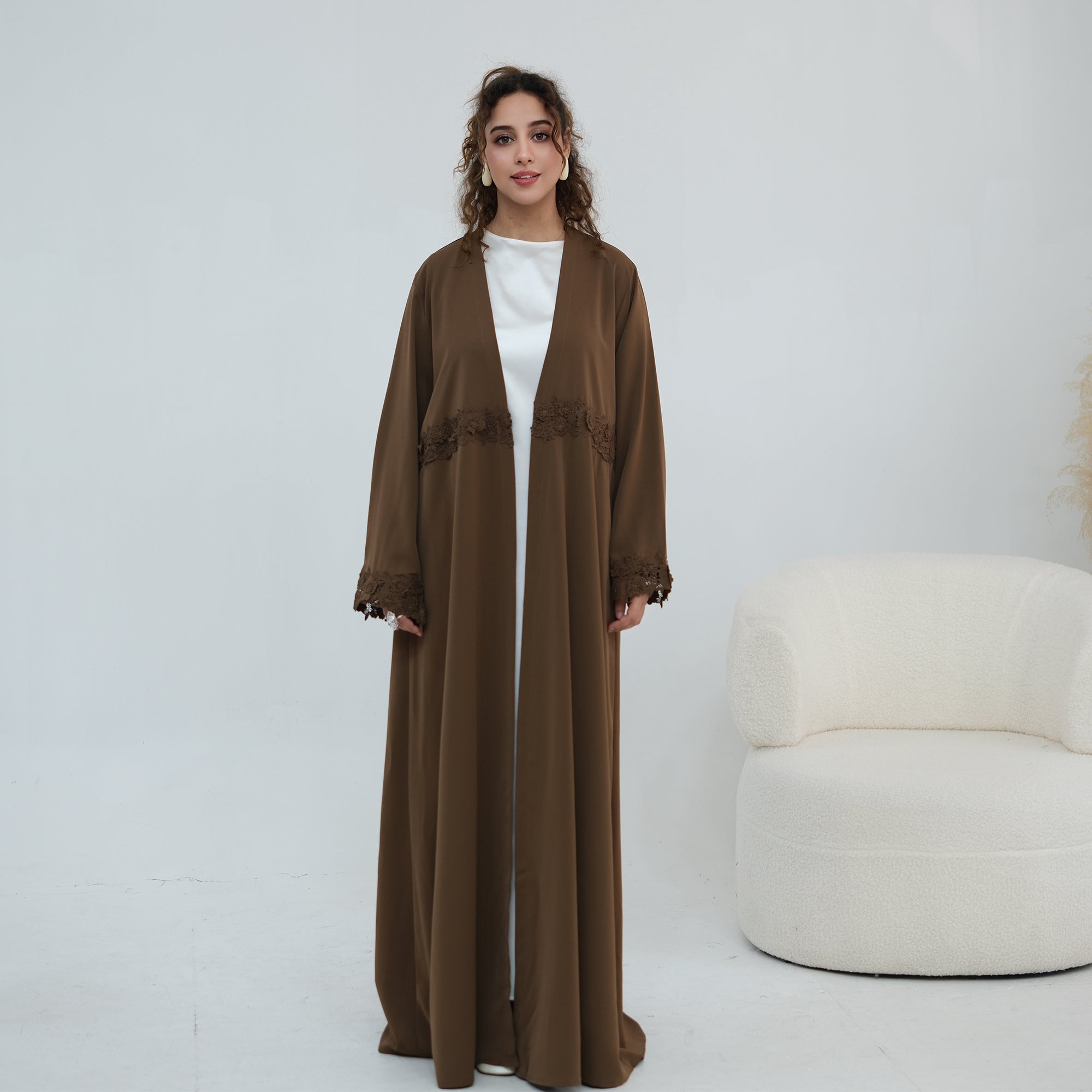 A woman stands in a studio wearing a long brown open-front abaya or kaftan with lace trim details, layered over a white top. A white curved armchair is visible to the right against a plain light background.