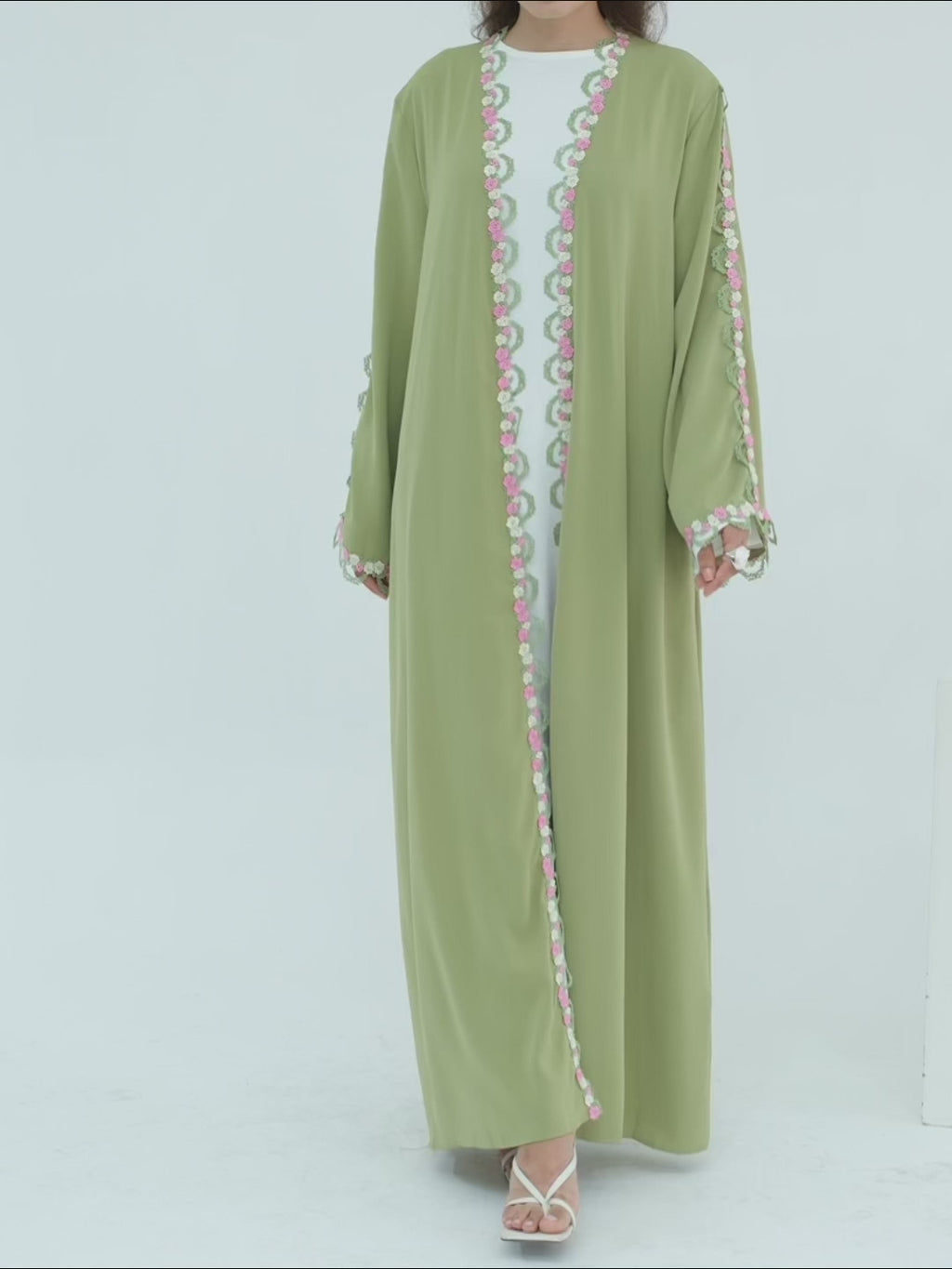 A woman modeling a sage green abaya with colorful embroidered details on the front opening, cuffs, and hem, paired with a white inner top and white sandals, set against a plain light gray background.