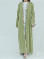 A woman modeling a sage green abaya with colorful embroidered details on the front opening, cuffs, and hem, paired with a white inner top and white sandals, set against a plain light gray background.