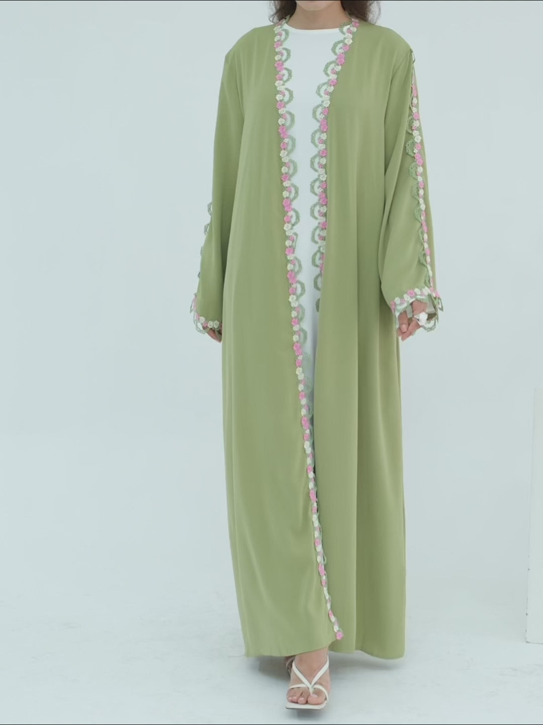 A woman modeling a sage green abaya with colorful embroidered details on the front opening, cuffs, and hem, paired with a white inner top and white sandals, set against a plain light gray background.