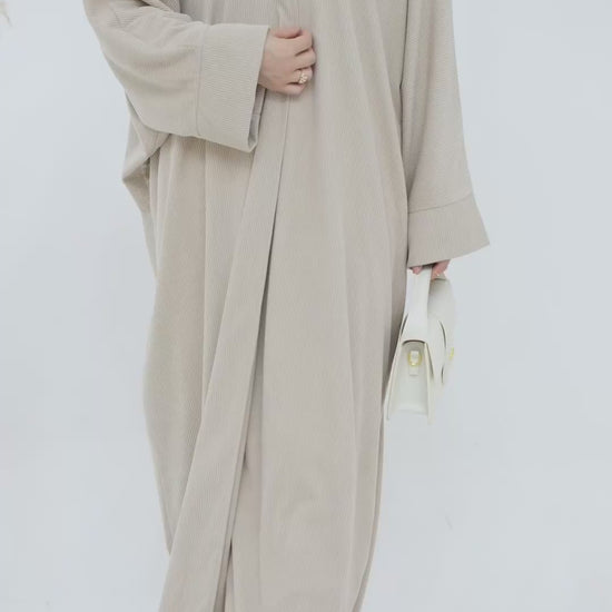 Person wearing a beige corduroy abaya with a white background and a vase with dried plants on the left. EverydayCorduroyAbaya_Beige1 - Noor & Grace