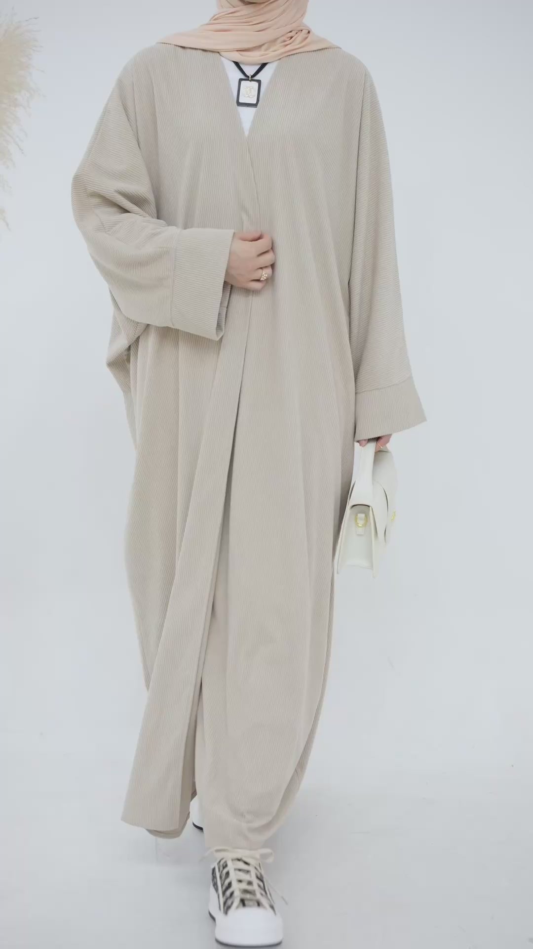 Person wearing a beige corduroy abaya with a white background and a vase with dried plants on the left. EverydayCorduroyAbaya_Beige1 - Noor & Grace