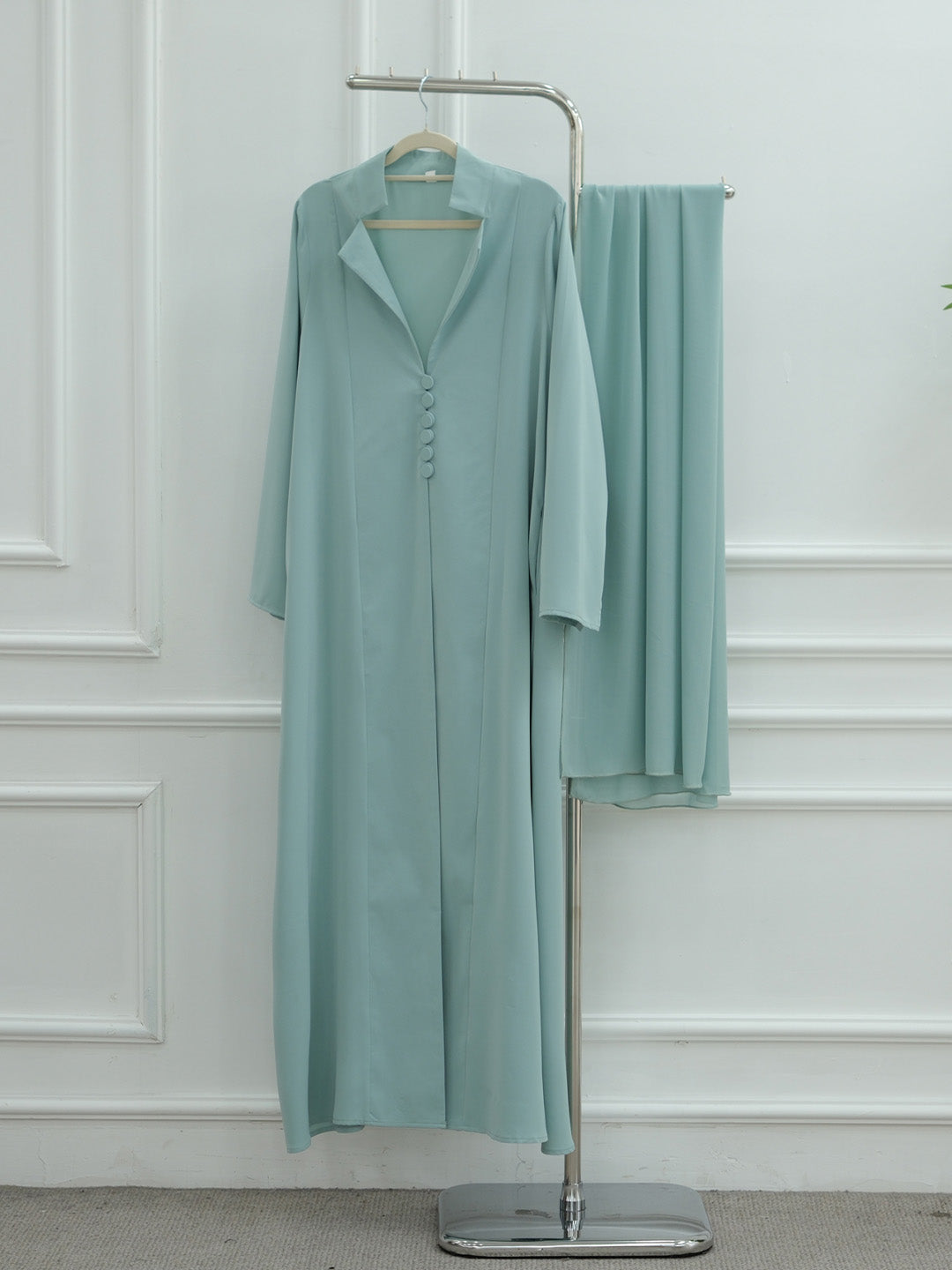 A woman viewed from behind wearing a flowing, long-sleeved abaya in a soft dusty blue-green color against a plain white studio background.