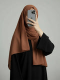 A woman wearing a draped brown hijab and a black abaya takes a mirror selfie with a gray smartphone against a plain light gray background.