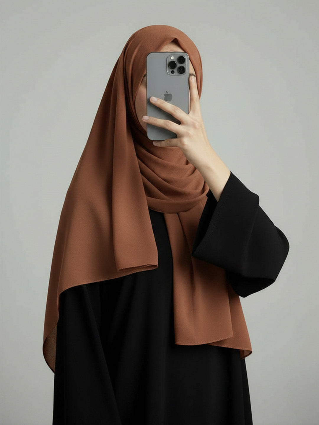 A woman wearing a draped brown hijab and a black abaya takes a mirror selfie with a gray smartphone against a plain light gray background.