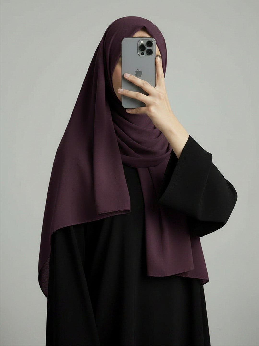 A woman wearing a deep plum purple hijab and a black abaya takes a mirror selfie with her phone against a solid gray background.