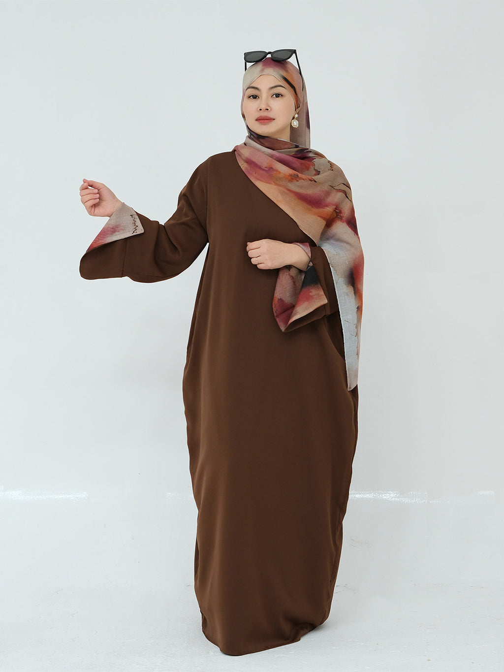 A woman modeling a long brown abaya dress with wide sleeves, paired with a patterned hijab in shades of pink, beige, and brown, against a plain white studio background.