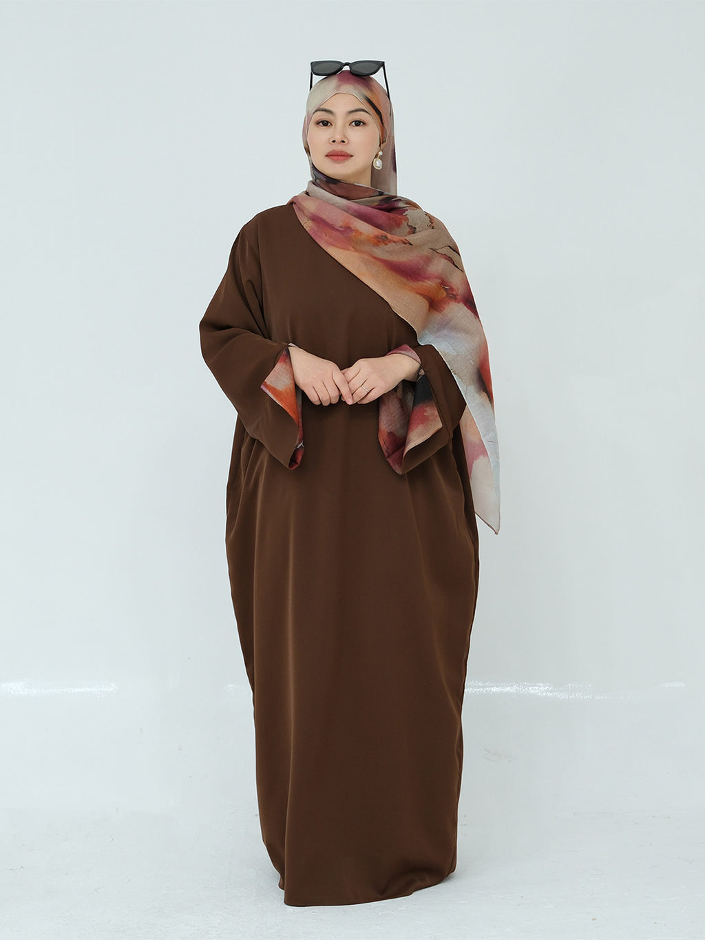 A woman modeling a long brown modest dress with wide sleeves, paired with a patterned brown, pink, and beige hijab and sunglasses resting on her head, set against a plain light gray studio background.