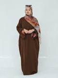 A woman modeling a long brown modest dress with wide sleeves, paired with a patterned brown, pink, and beige hijab and sunglasses resting on her head, set against a plain light gray studio background.