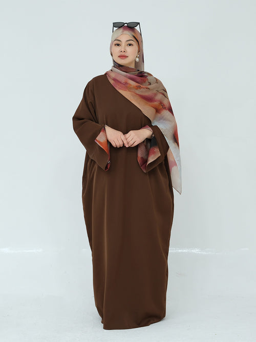 A woman modeling a long brown modest dress with wide sleeves, paired with a patterned brown, pink, and beige hijab and sunglasses resting on her head, set against a plain light gray studio background.