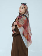 A woman wearing a brown dress and a large printed hijab scarf with an abstract watercolor pattern in pink, orange, and gray, posed against a light gray studio background.