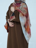 A person wearing a brown abaya and a large abstract-print hijab scarf in shades of pink, orange, beige, and dark brown, posed against a light gray studio background.