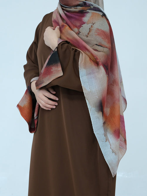 A person wearing a brown abaya and a large abstract-print hijab scarf in shades of pink, orange, beige, and dark brown, posed against a light gray studio background.