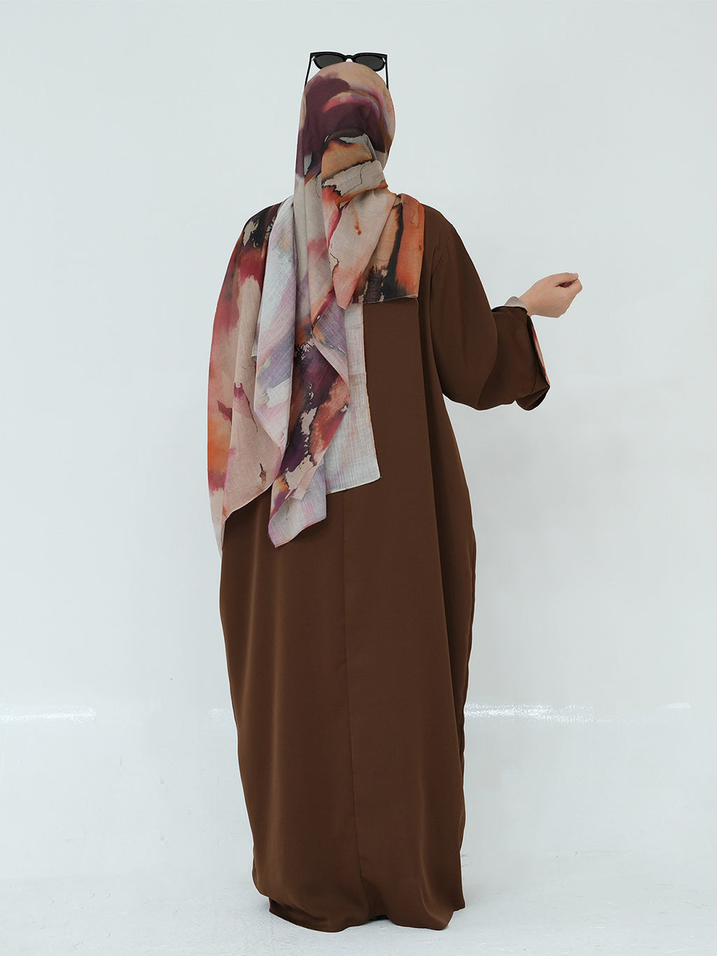 A person wearing a long brown abaya dress with a matching solid brown headscarf and a patterned hijab draped over the shoulders, standing against a plain light gray studio background.
