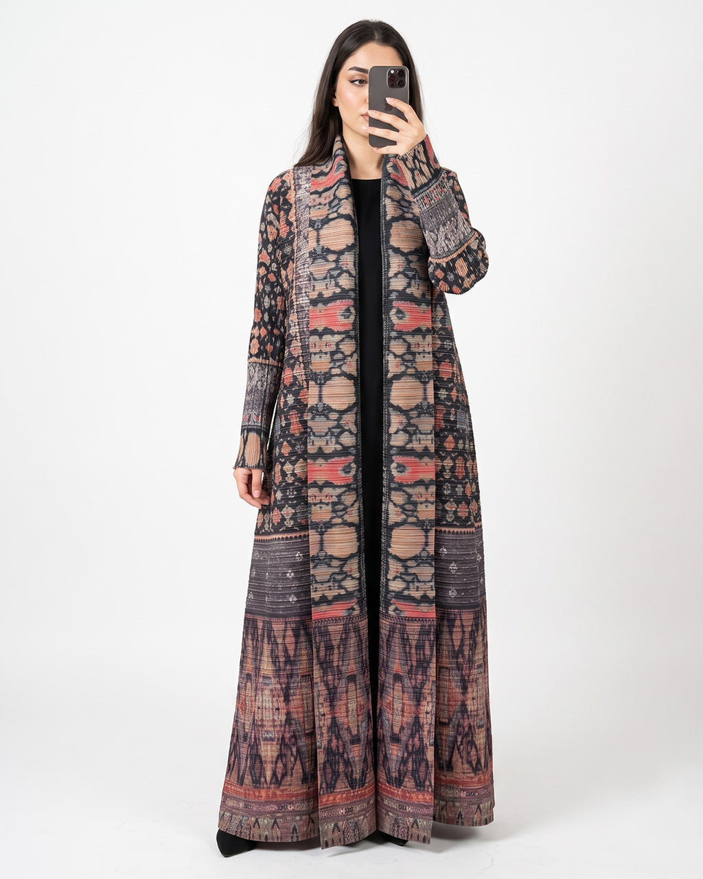 A woman takes a mirror selfie wearing a long, open-front maxi dress with an intricate multicolor geometric and tribal pattern in shades of brown, beige, red, and black.