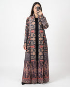 A woman takes a mirror selfie wearing a long, open-front maxi dress with an intricate multicolor geometric and tribal pattern in shades of brown, beige, red, and black.