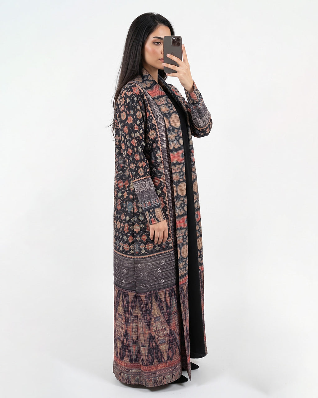 A woman takes a mirror selfie wearing a long, open-front kaftan-style coat with an intricate multicolor floral and geometric pattern in dark tones, over a black inner dress, against a plain white studio background.