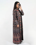 A woman takes a mirror selfie wearing a long, open-front kaftan-style coat with an intricate multicolor floral and geometric pattern in dark tones, over a black inner dress, against a plain white studio background.