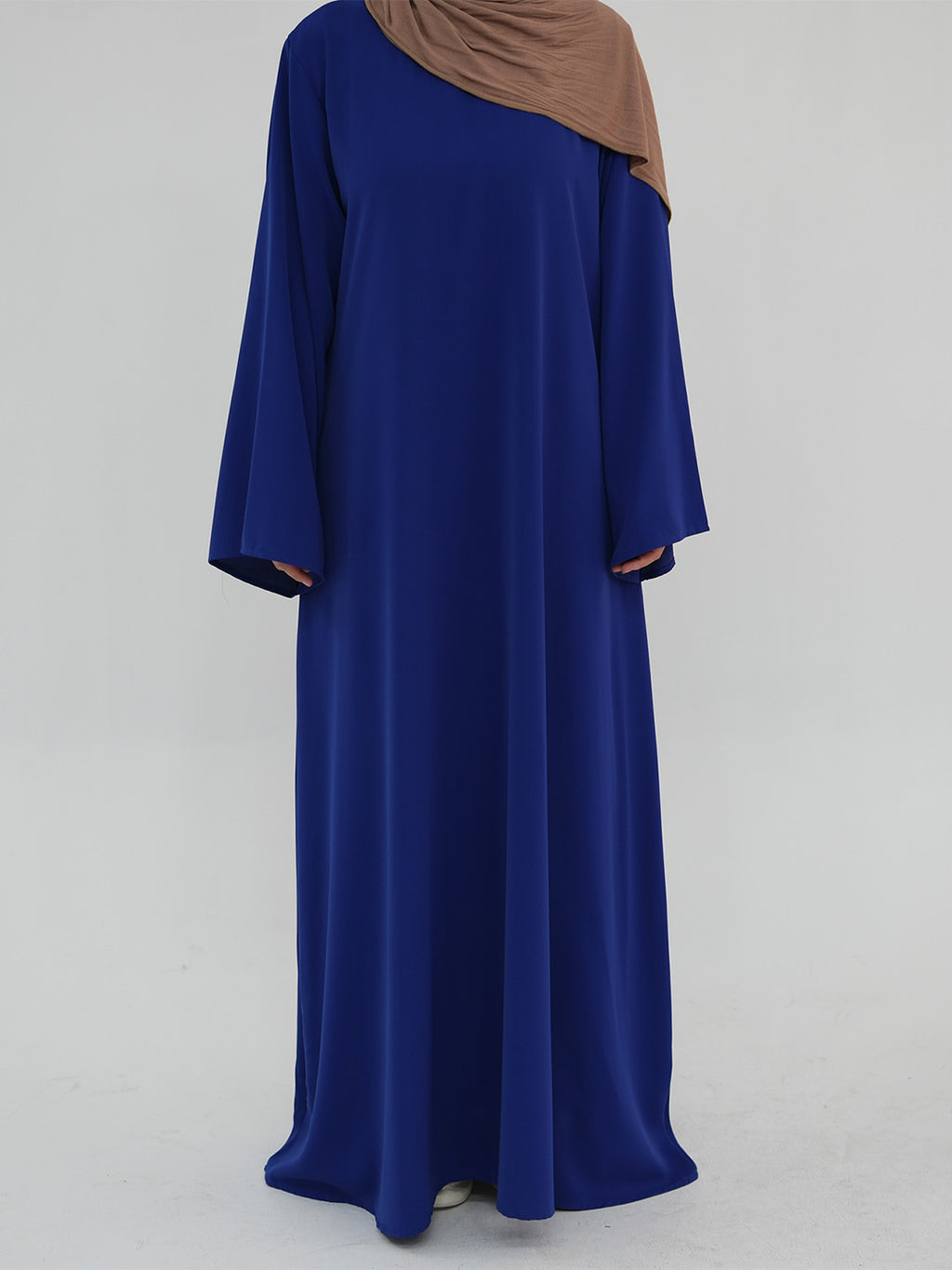 A woman wearing a long, flowing royal blue abaya dress with a draped brown hijab, photographed against a plain light gray studio background.
