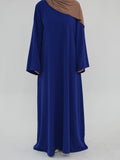 A woman wearing a long, flowing royal blue abaya dress with a draped brown hijab, photographed against a plain light gray studio background.