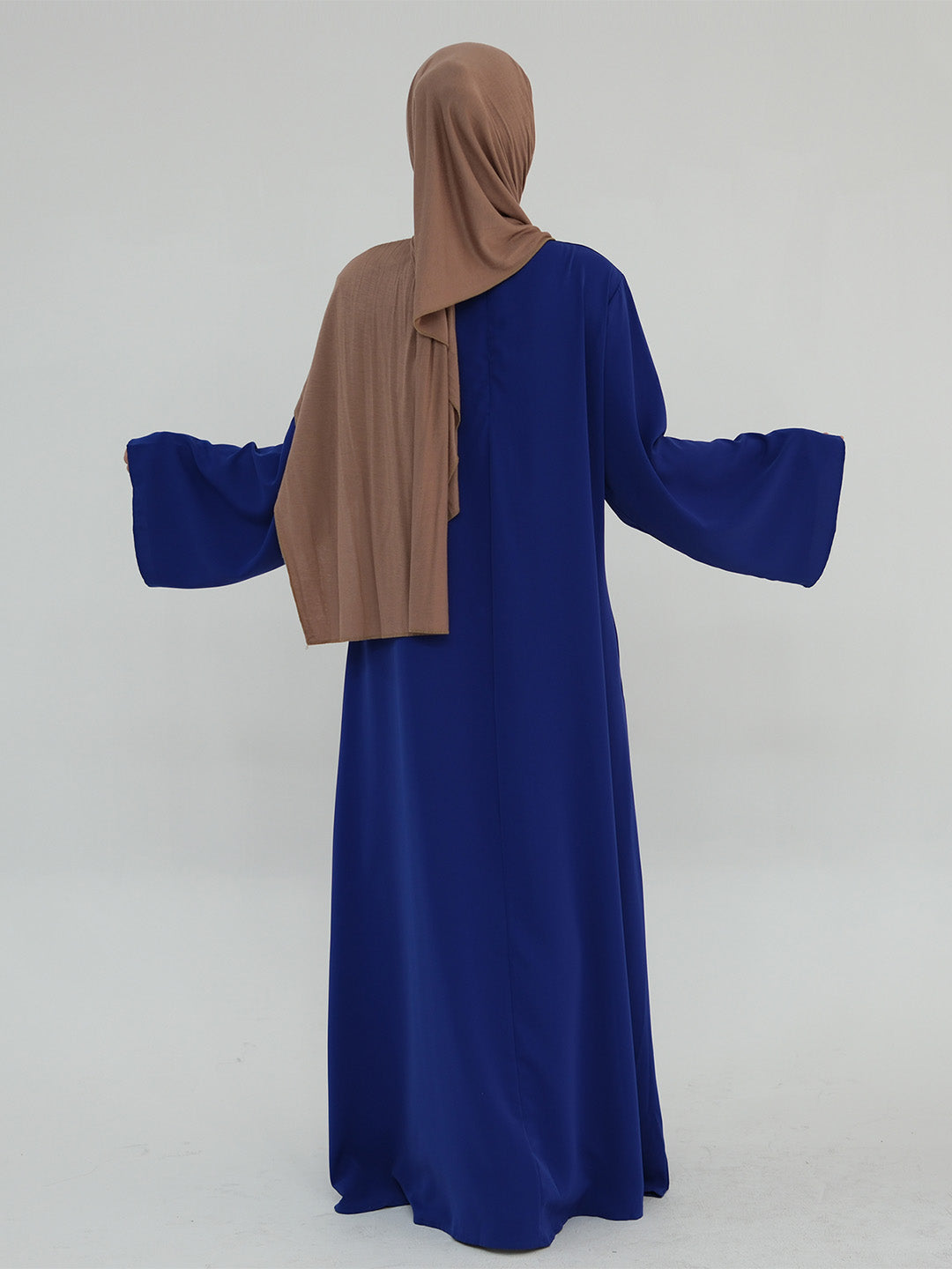 A woman shown from the back wearing a long royal blue abaya with wide bell sleeves and a taupe-brown hijab, photographed against a plain light gray studio background.