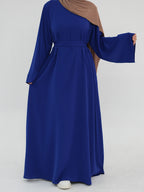 A woman wearing a long-sleeved royal blue abaya dress with a matching waist tie and a brown hijab, standing against a light gray studio background.