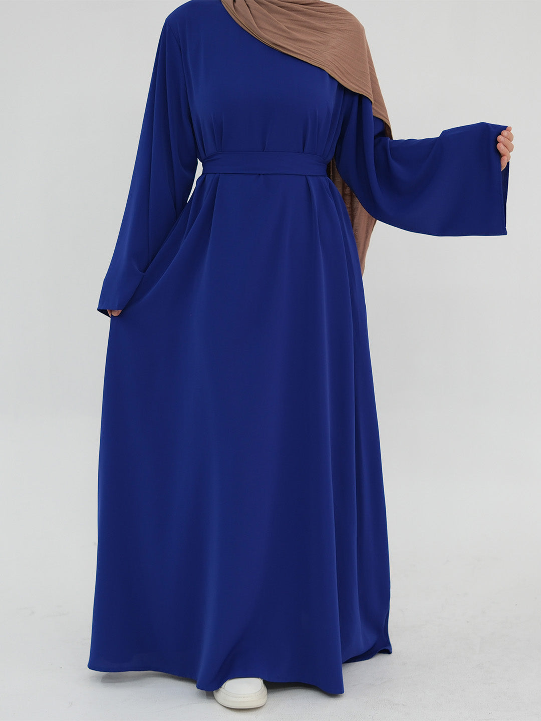 A woman wearing a long-sleeved royal blue abaya dress with a matching waist tie and a brown hijab, standing against a light gray studio background.