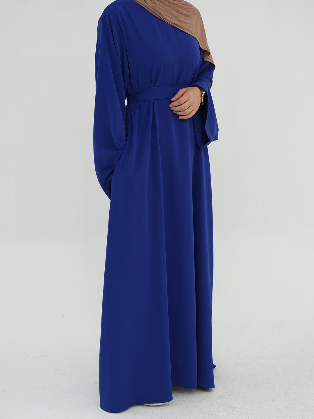A woman wearing a long-sleeved royal blue maxi abaya dress with a matching waist tie and a brown hijab, shown from the side against a plain light gray background.