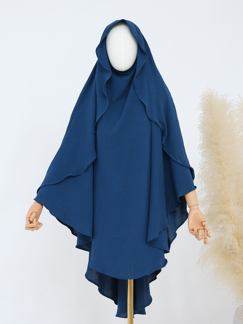 A navy blue draped hijab or modest garment displayed on a mannequin with visible hands and a white mannequin head, against a light gray background with a beige decorative pampas grass element at the right.