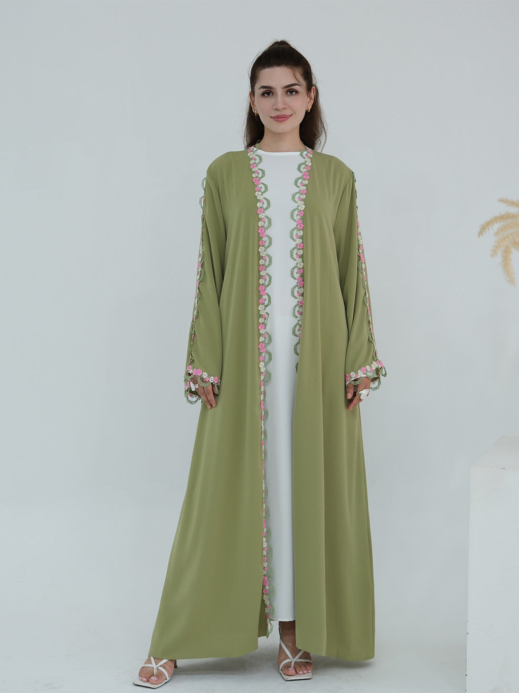 A woman modeling a long olive green abaya dress with pink and green embroidered patterns along the front opening and cuffs, worn over a white inner dress.