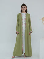 A woman modeling a long olive green abaya dress with pink and green embroidered patterns along the front opening and cuffs, worn over a white inner dress.