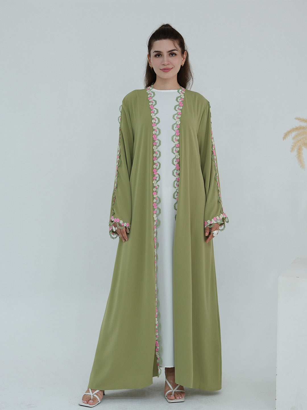 A woman modeling a long olive green abaya dress with pink and green embroidered patterns along the front opening and cuffs, worn over a white inner dress.