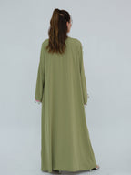 A woman viewed from behind wearing a long sage green abaya dress with white lace trim at the cuffs, set against a plain light gray studio background.