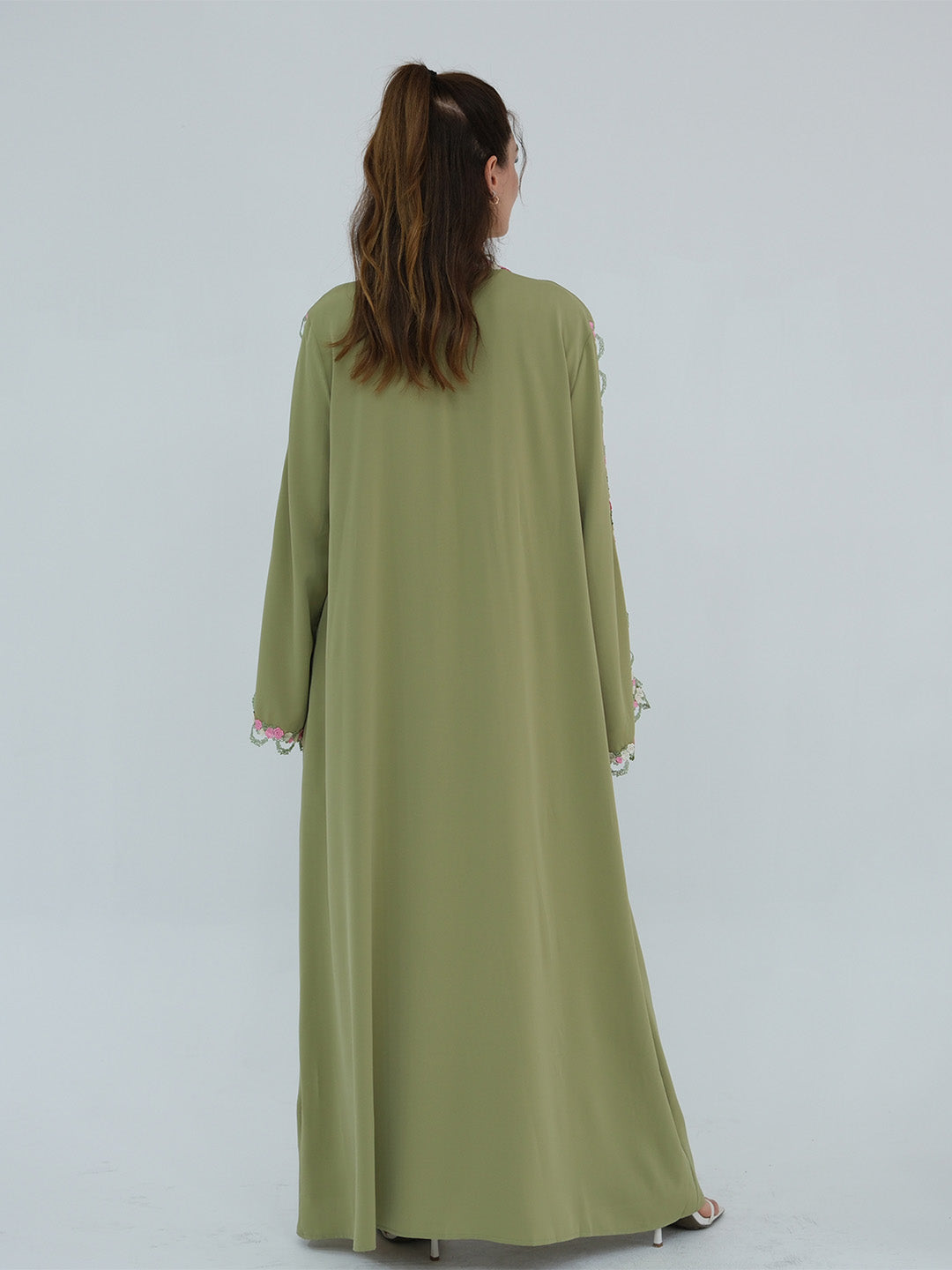A woman viewed from behind wearing a long sage green abaya dress with white lace trim at the cuffs, set against a plain light gray studio background.