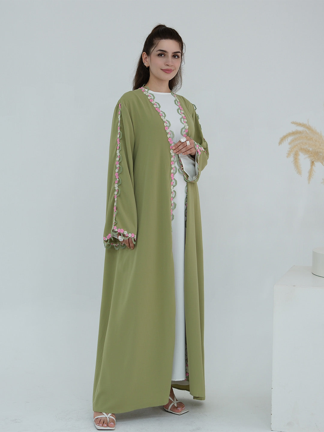 A woman modeling a long sage green abaya with pink and white floral embroidery on the front opening and sleeve cuffs, worn over a white dress in a studio setting.