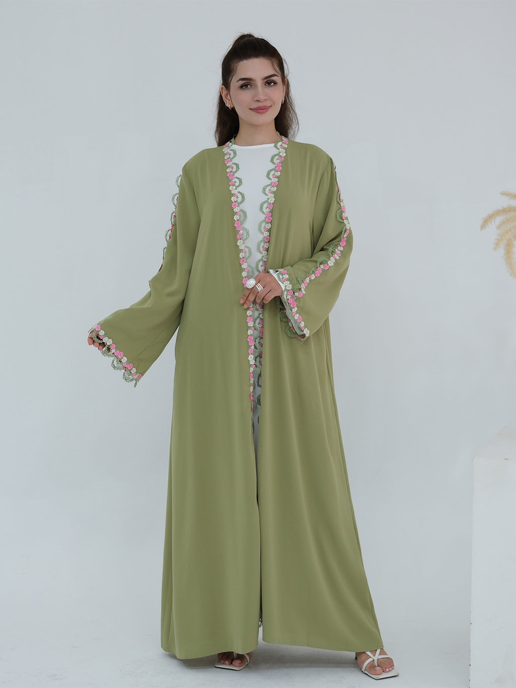 A woman modeling a sage green abaya with colorful embroidered details on the front opening, cuffs, and hem, paired with a white inner top and white sandals, set against a plain light gray background.