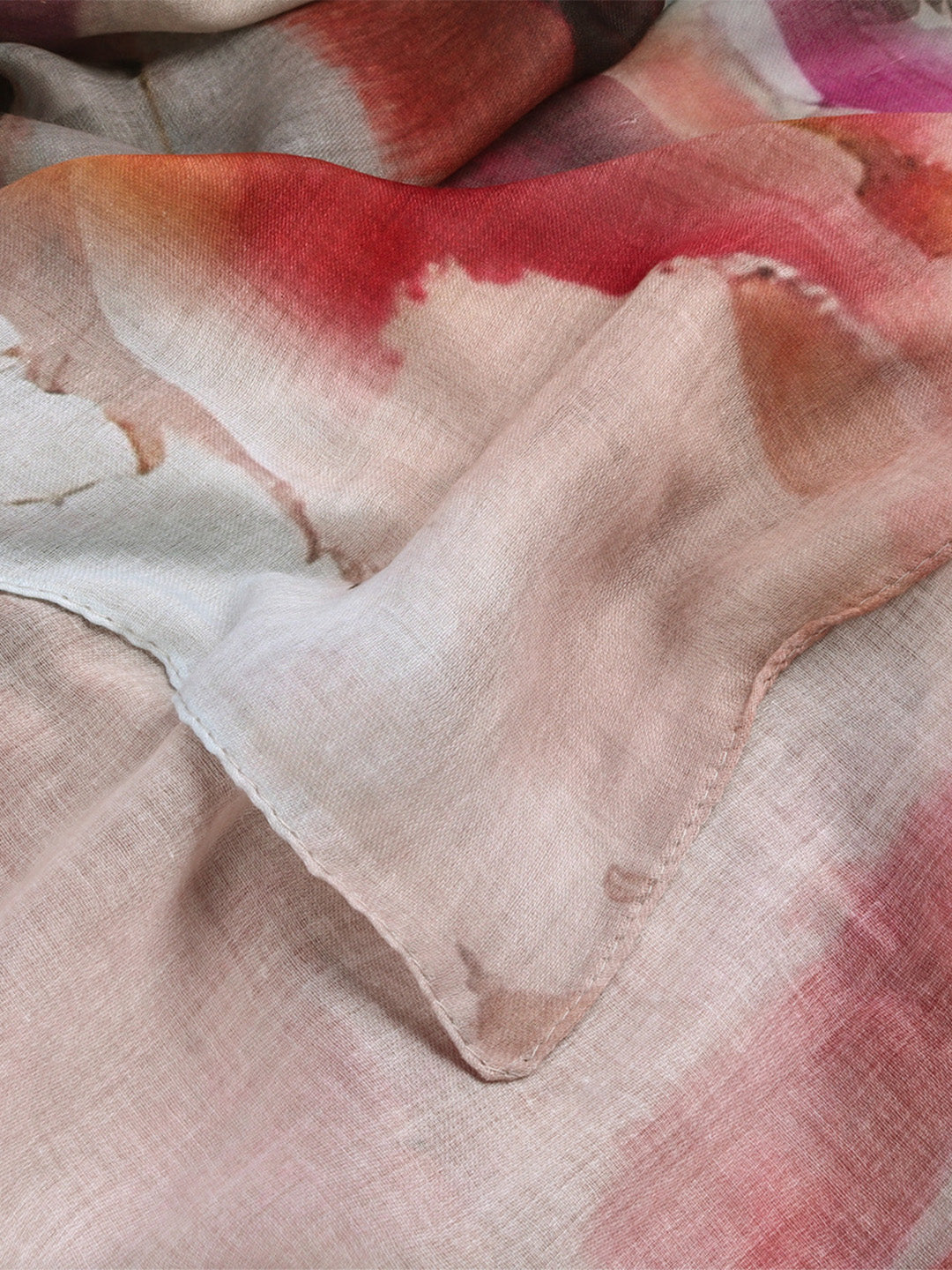 A close-up of a lightweight fabric with a soft tie-dye or watercolor pattern in shades of pink, red, beige, and white.