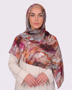 A woman wearing a vibrant printed modal hijab with an intricate multi-color marble pattern featuring orange, purple, and charcoal grey tones, paired with a cream outfit