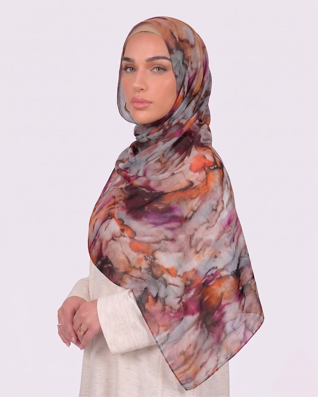 A woman wearing a vibrant printed modal hijab with an intricate multi-color marble pattern featuring orange, purple, and charcoal grey tones, paired with a cream outfit